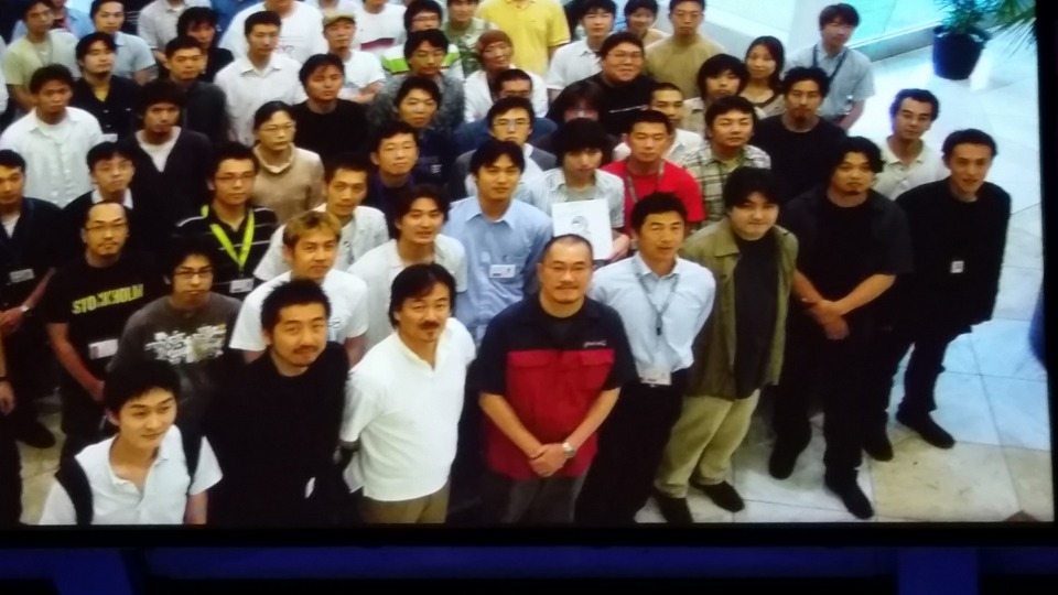 Photo of a projection of a photo of Sakaguchi posing with the Blue Dragon development team.